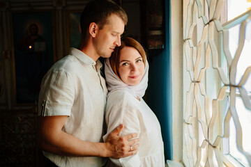 man and woman in headscarf and light-colored robes by window in church. 