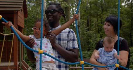 A father helps his daughter overcome an obstacle made of ropes in a playground. Communication between adults on the topic of raising children.