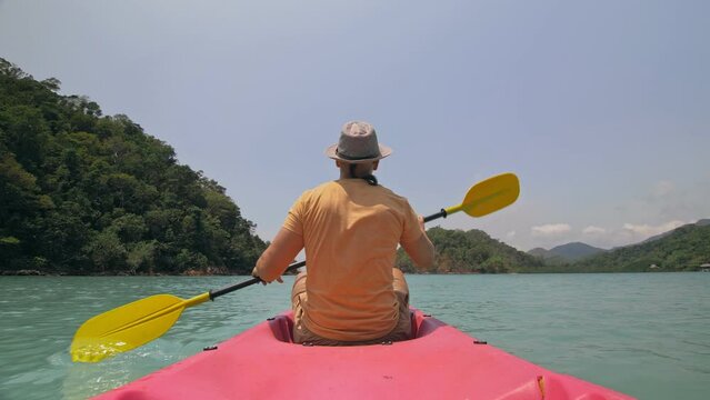 Man with sunglasses and hat rows pink plastic canoe along sea against green hilly islands with wild jungles. Traveling to tropical countries.