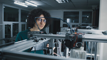 Young man in glasses with curly hair checking 3D printer with plastic model and using tablet during work in modern workshop