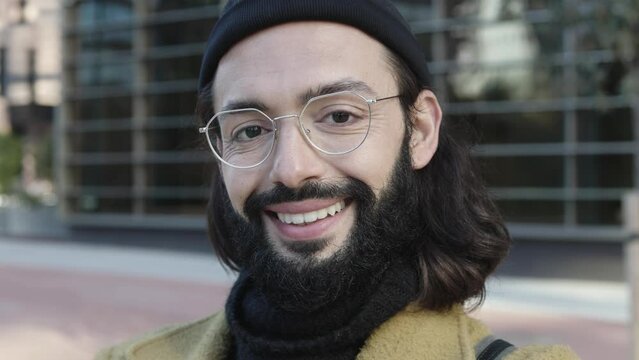 Portrait of positive young hipster man smiling at camera - Handsome trendy bearded guy with hat and glasses standing outdoors - Fashion and trendy business people concept. High quality 4k footage