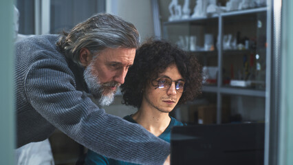 Mature bearded man in casual clothes supervising young employee creating 3D model on computer...