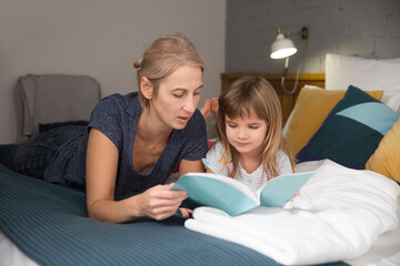 mom and little daughter read a book lying in the bedroom on the bed
