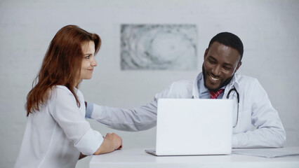 cheerful african american doctor talking with redhead patient near laptop on desk.