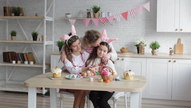 an adult mother hugs and gently kisses her daughters, helps to paint Easter eggs in the home kitchen. preparation for the Easter holiday family traditions