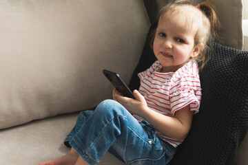 Little girl is sitting on sofa with smart phone and sticking her tongue.