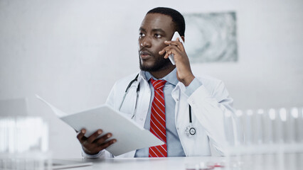 serious african american doctor talking on smartphone and holding paper folder in clinic.