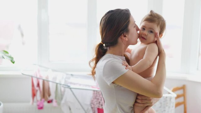 satisfied young mom kisses and hugs her baby at big window at home. happy mother kissing adorable baby. Concept of family happiness and child development