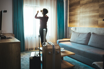Fototapeta na wymiar Luggage in modern hotel room with happy young adult female relaxing nearly window, African woman tourist looking to beautiful nature view. 