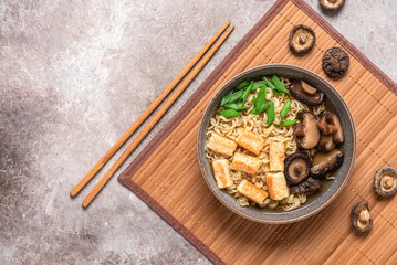 Asian vegan ramen soup with noodles, roasted tofu cheese and shiitake mushrooms in a bowl on a bamboo mat. Gray concrete grunge background. Top view.