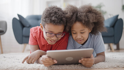 Cheerful black siblings lying on floor with tablet, having fun together, smiling