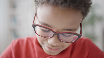 Cute african american boy in eyeglasses looking down, reading book, writing down homework or drawing picture sitting at table, hobby