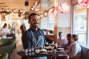 Portrait of smiling waiter with food while customer sitting in background at bar