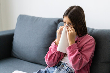 Little girl sick blowing her nose with tissue and cover with blanket sitting on sofa in the living room at home.