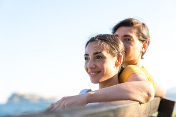 Handsome man wearing yellow tshirt and blue swimsuit sits with beautiful woman in dungarees in a park in a sunny day during holidays. Young adults couple sitting togheter surrounded by green trees