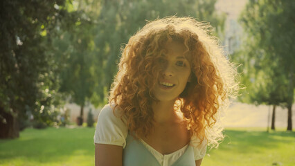 sunshine on curly red hair of woman smiling while looking at camera in park.