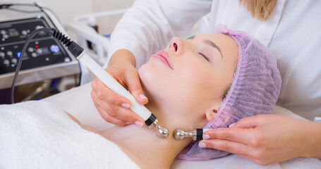 Obraz na płótnie Canvas Woman in a spa salon on cosmetic procedures for facial care. Cosmetologist making a woman a therapeutic procedure on a face. Beautician makes medical procedures using a professional equipment.