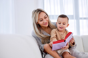 friendly family young mother babysitter hold read book relax embrace cute little child boy, smiling parent mum tell small kid funny fairy tale story sit on sofa having fun together at home