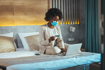 A young woman buys online at hotel, holds a credit card, pays on the web via a laptop, makes secure online payments, buys goods in an online store, checks her account balance, wears a protective mask