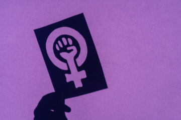 Shadow of the symbol of the fight for feminism on a purple background, clenched fist of a woman in...