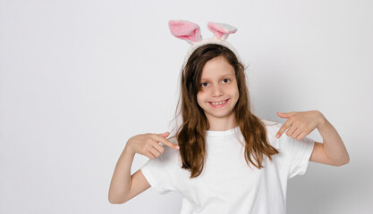 Cute dark-haired girl 9 years old. With bunny ears on the head. Pointing at a white T-shirt. Easter...