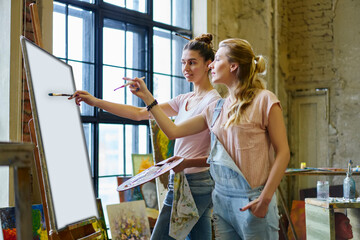 Caucasian women in casual clothes discussing art technique while drawing picture in loft interior, talented teacher and student talking about canvas on easel collaborate in workshop studio