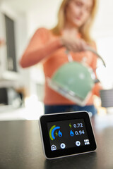Close Up Of Smart Energy Meter In Kitchen Measuring Electricity And Gas Use With Woman Boiling...