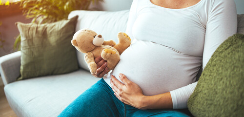 Pregnant Woman Holding Teddy Bear and smiling, sitting on the floor in her living room. Wearing...
