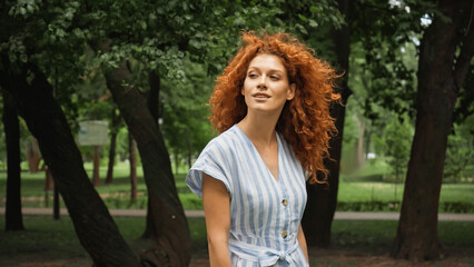 pleased young woman with red hair standing in blue striped dress in green park.