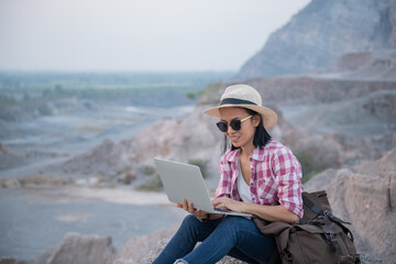 Young woman sitting with laptop on boulder against mountains. female working on laptop sitting on the peak of a mountain at sunset. Technology, internet, staying online.