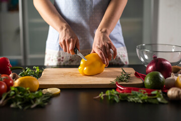 Hands of Young Woman Slicing Fresh Yellow Pepper on the Wooden Cutting Board with Knife in the Kitchen. Making Natural Vegan Healthy Salad
