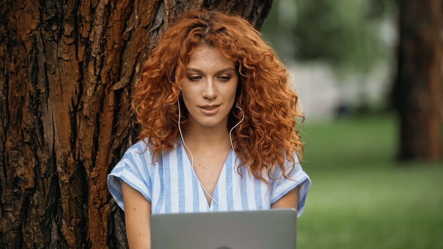 young redhead woman listening music in earphones while using laptop in park.