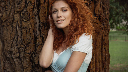 young and curly redhead woman smiling near tree in park.