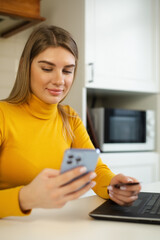 Fototapeta premium Happy white woman paying with credit card. Young female shopping online with modern smart phone and bank card. Cheerful blonde making money transfer in mobile phone app with smile