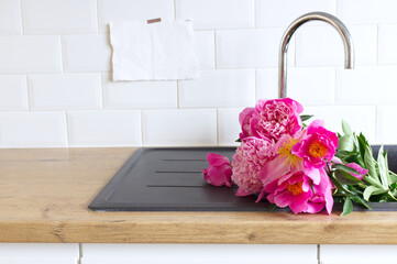 Closeup of kitchen interior. White brick wall, metro tiles, wooden countertops with a chopping board. Pink peonies in the black sink. Modern Scandinavian design. Home staging, cleaning concept.