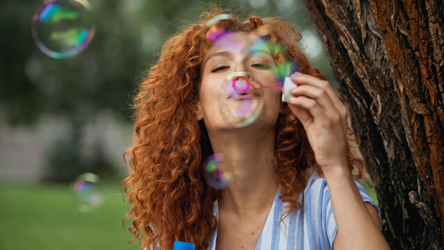 cheerful redhead woman blowing soap bubbles in park.