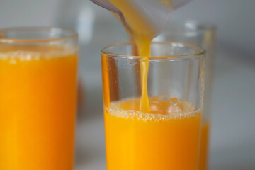 Woman is pouring fresh orange juice from electric juicer into three glasses, closeup view. Healthy vegan ripe fruit juice, natural vitamins from food. Vegan healthy organic nutrition.