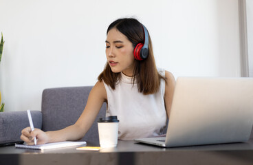 Asian woman working on laptop at home having an online meeting and writing notes in a notepad.