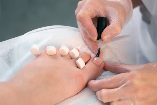 Pedicurist master is applying transparent base coal for shellac on woman's nails on toes, closeup hands. Preparing nails for shellac in beauty salon. Hygiene and care for feet.
