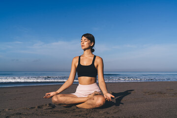 Fototapeta na wymiar Caucasian female with closed eyes practicing yoga at coastline reaching healthy lifestyle and wellness, calm woman with flexible body sitting in lotus pose enjoying asana time for mindfulness