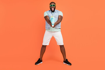 Photo of funny impressed guy dressed jeans waistcoat dark glasses shooting water gun isolated orange color background