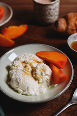 Homemade ricotta cheese or cheese curd served with fresh persimmon and honey and croissante on wooden background. Healthy breakfast. Haze effect. Selective focus