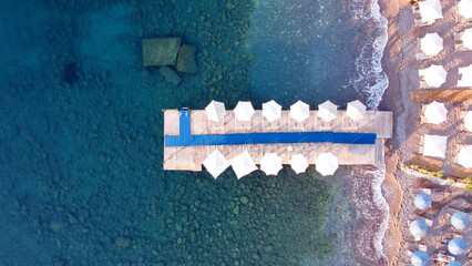 Top view of wooden Pier with beach umbrellas and sunbeds in touristic resort. Transparent sea water. Rocks on sea bottom. Calm sea by drone view
