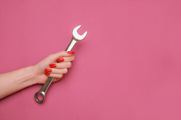 A steel wrench in a female hand with red manicure on a pink background, copy-space.