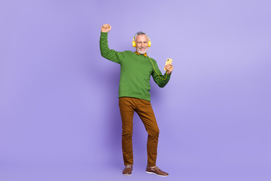 Photo of lucky retired man wear green sweater enjoying songs modern device rising fists isolated violet color background