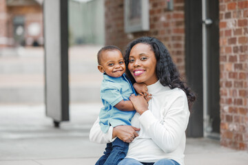 A beautiful African-American mom sitting on steps outdoors and hugging her toddler age son and they...