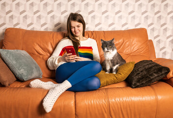 Young cute girl in glasses wearing white sweater with rainbow art and tight blue leggins sitting on orange leather sofa at home with her gray female cat working remotely with smartphone