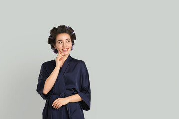 Happy young woman in silk bathrobe with hair curlers on grey background, space for text