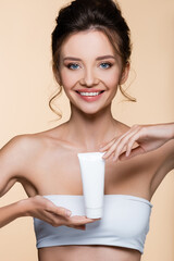 Obraz na płótnie Canvas Smiling brunette woman in white top holding cosmetic lotion isolated on beige.