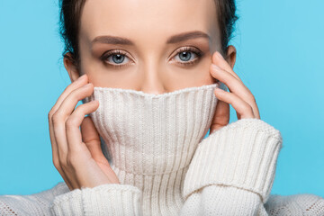 Young woman covering face with warm sweater isolated on blue.